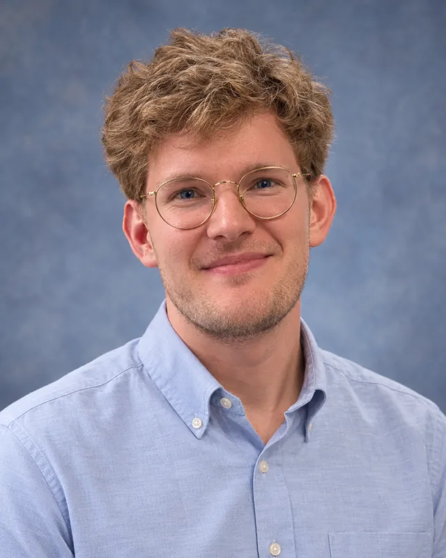 Official portrait of Ben Byler in a light blue shirt against a blue-gray studio backdrop